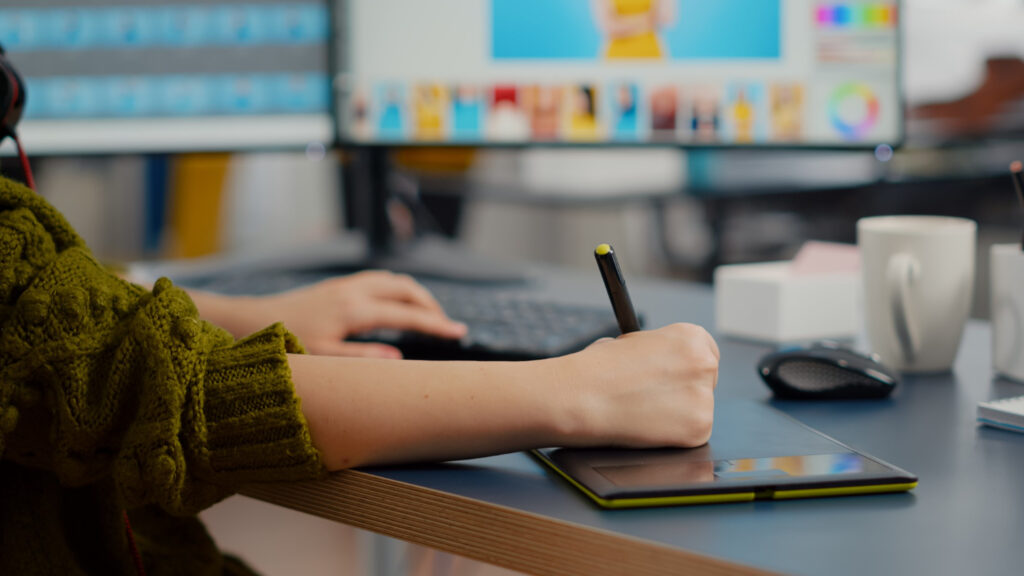 Close-up of designer using a pen tablet and keyboard while editing photos on a computer with dual monitors.