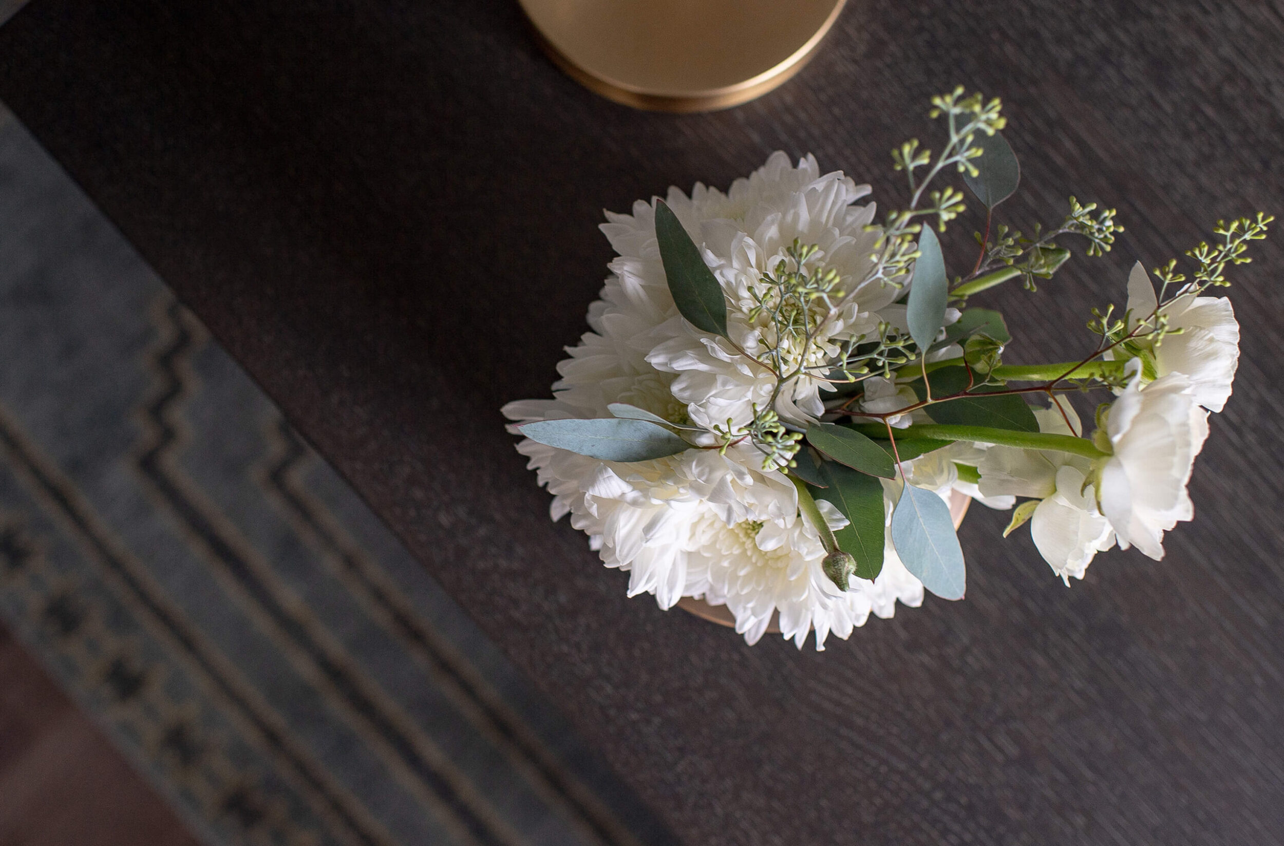 Overhead view of white floral arrangement with greenery in gold vase on dark wood desk with patterned rug below.