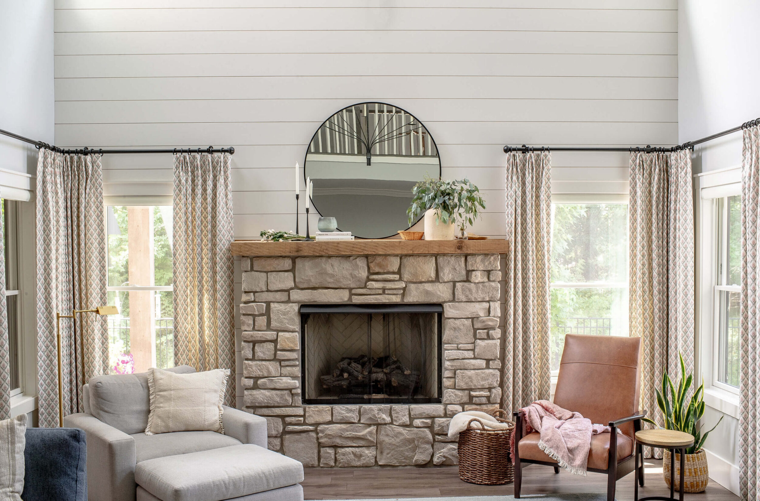 Living room fireplace view with stone surround, wood mantel, round mirror, and cozy seating on both sides.