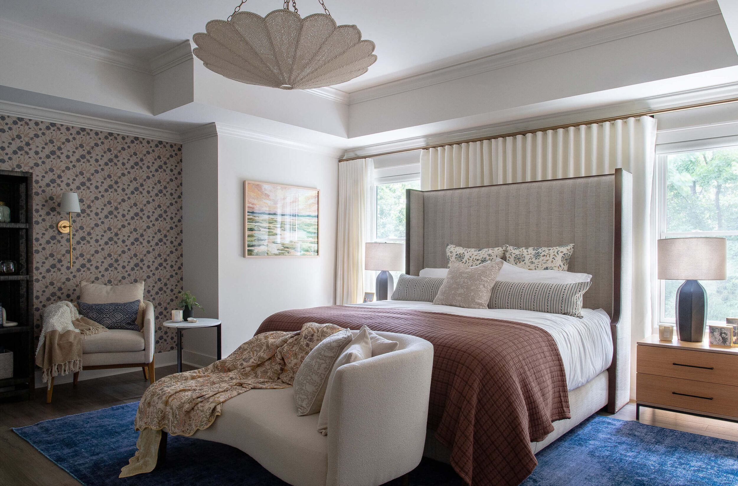 Elegant bedroom view with tall headboard, layered bedding, armchair corner, floral artwork, and soft ambient lighting.