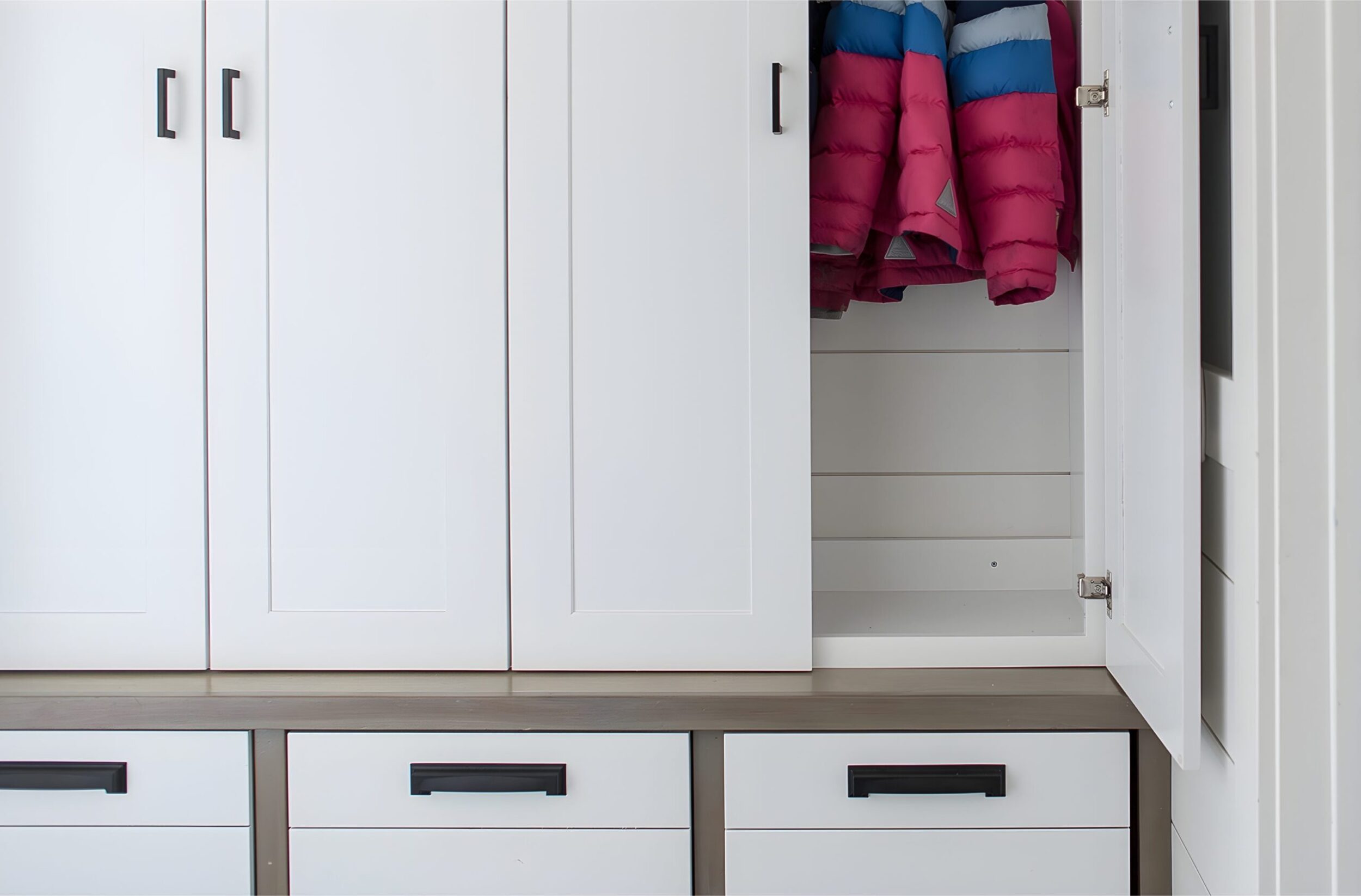 Tall white storage cabinets with black handles, partially open to reveal colorful winter jackets in a mudroom.