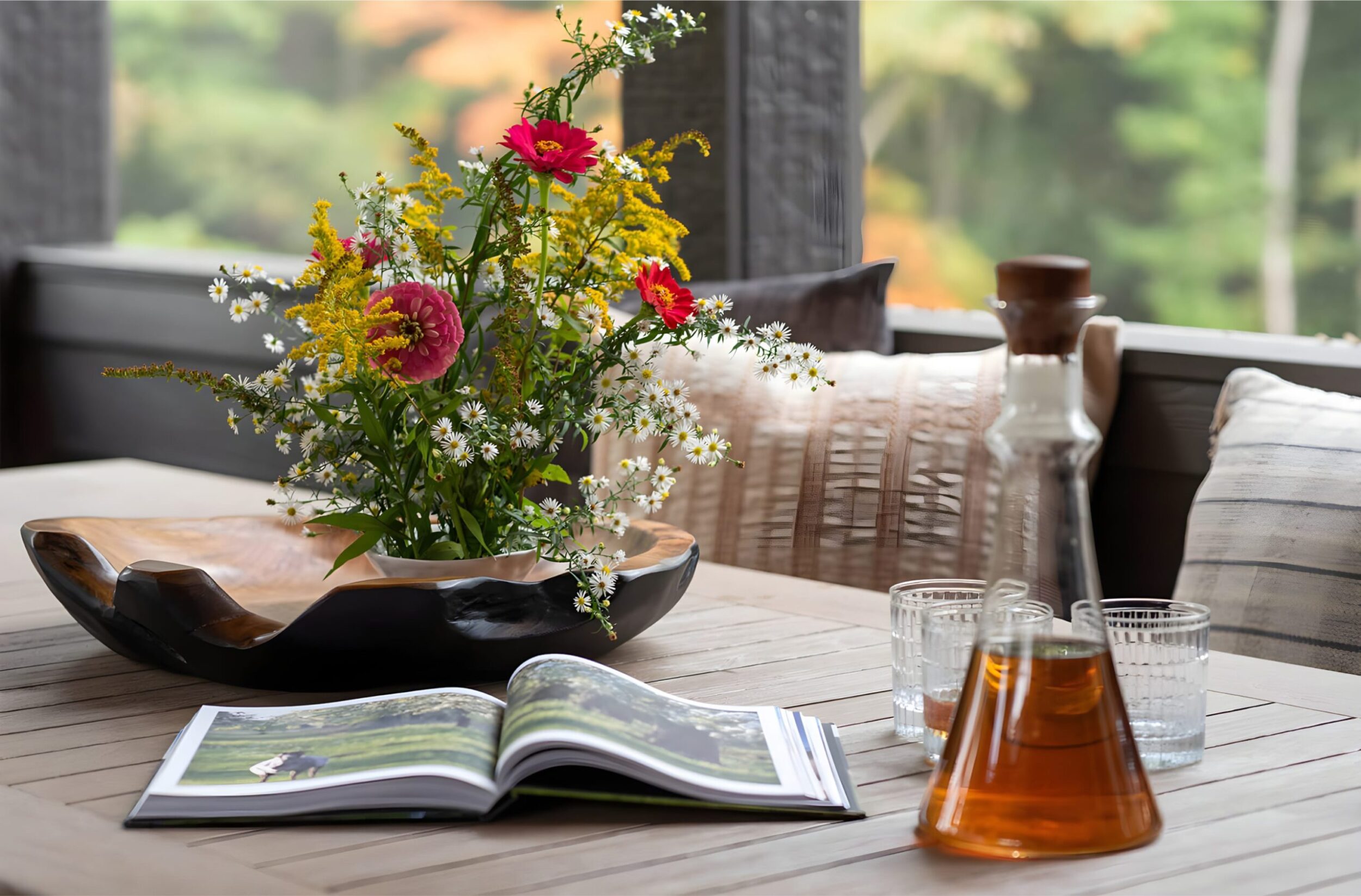 Wooden table with wildflower bouquet in black bowl, open book, glass decanter of tea, and glasses in cozy porch setting