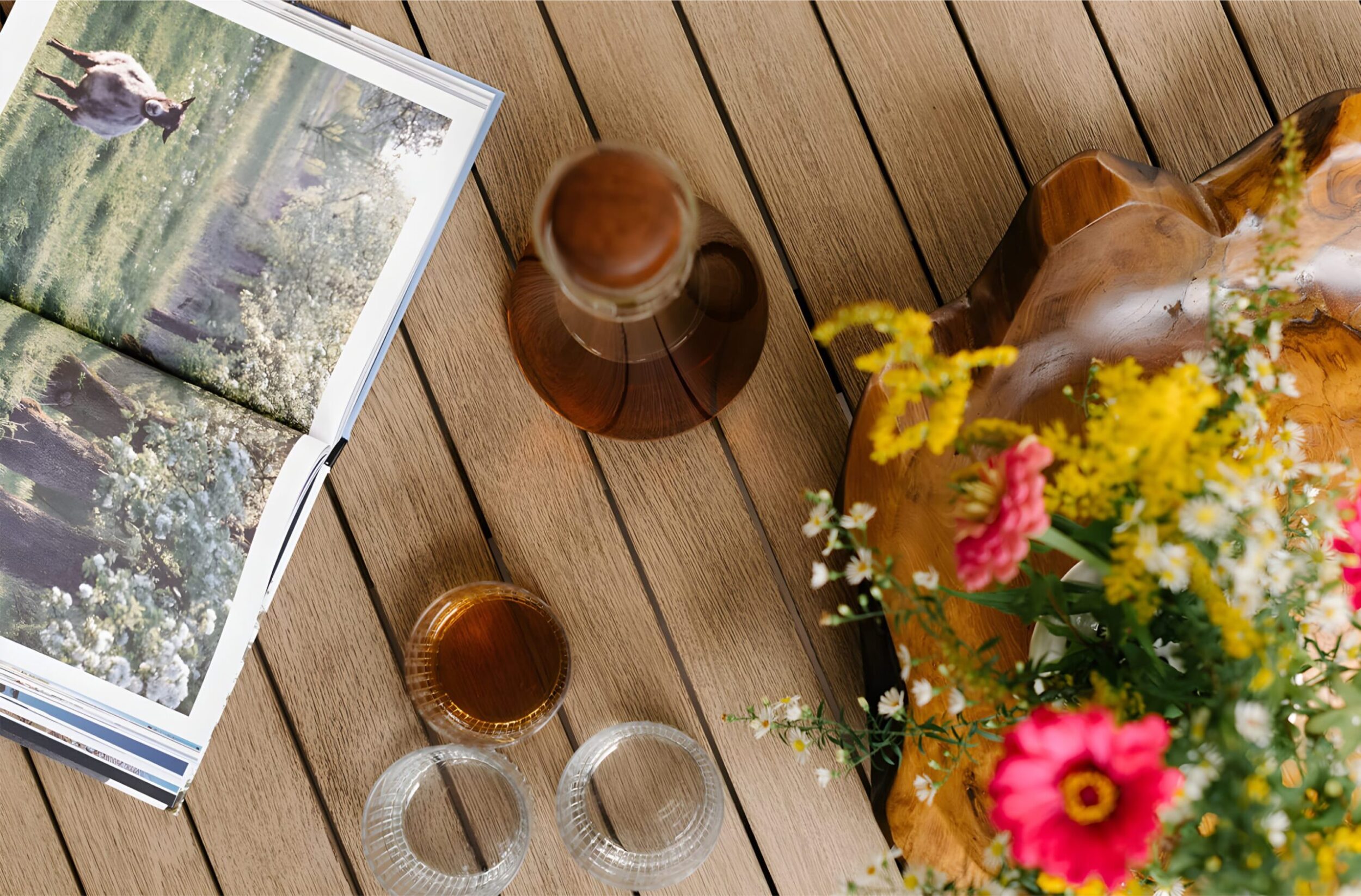 Overhead view of wooden table with open photo book, glass decanter of tea, wildflowers in wood bowl, and clear glass cups