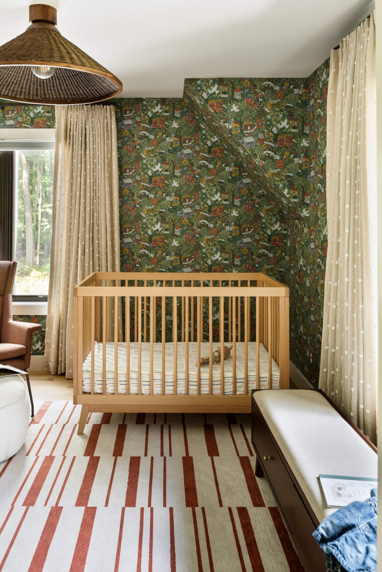Light wood crib on patterned rug in nursery with detailed green botanical wallpaper, beige dotted drapes, wicker pendant lamp.