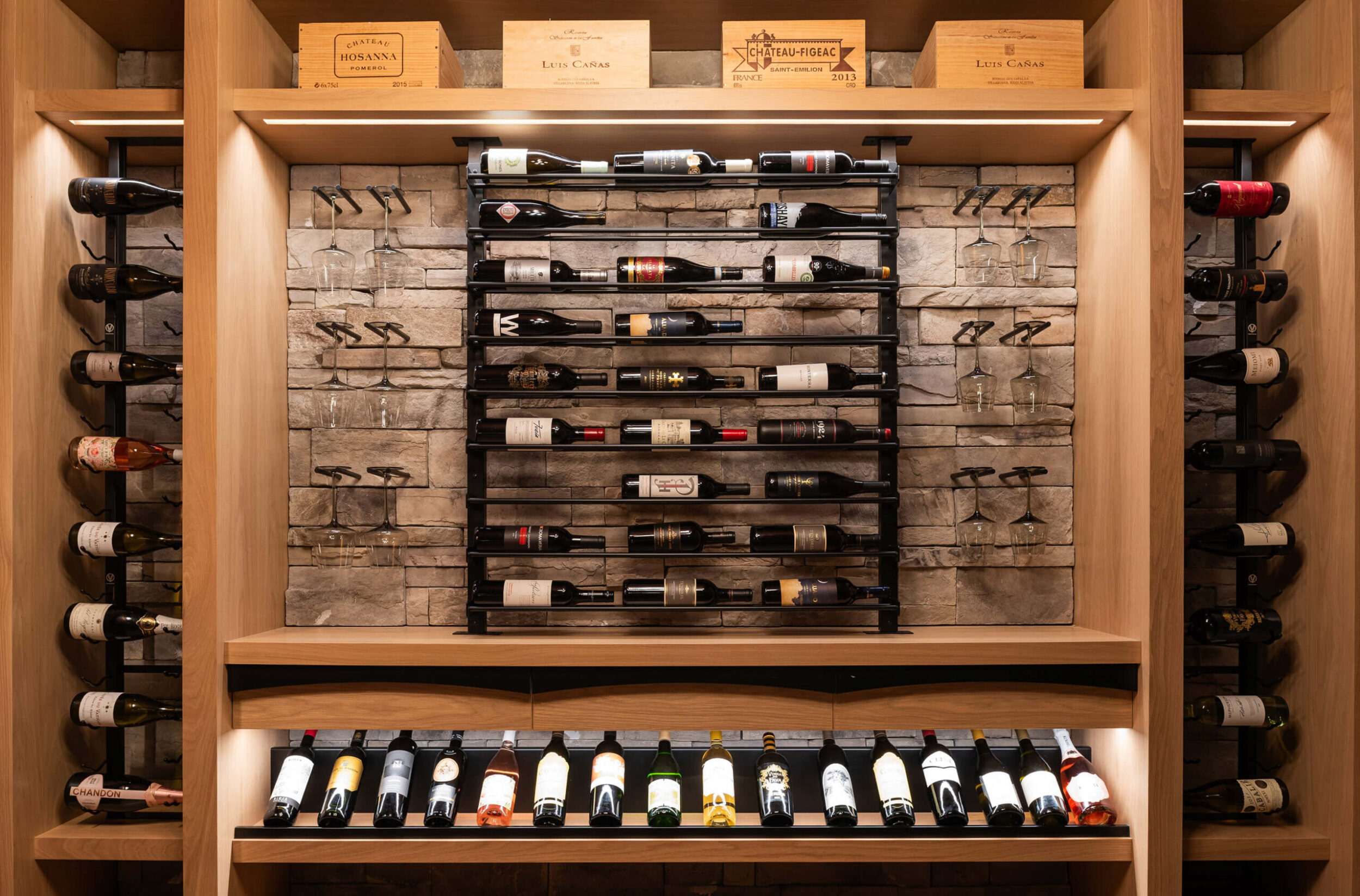 Wine cellar wall with stone backdrop, black racks holding assorted wine bottles, glass holders, and wooden shelves above