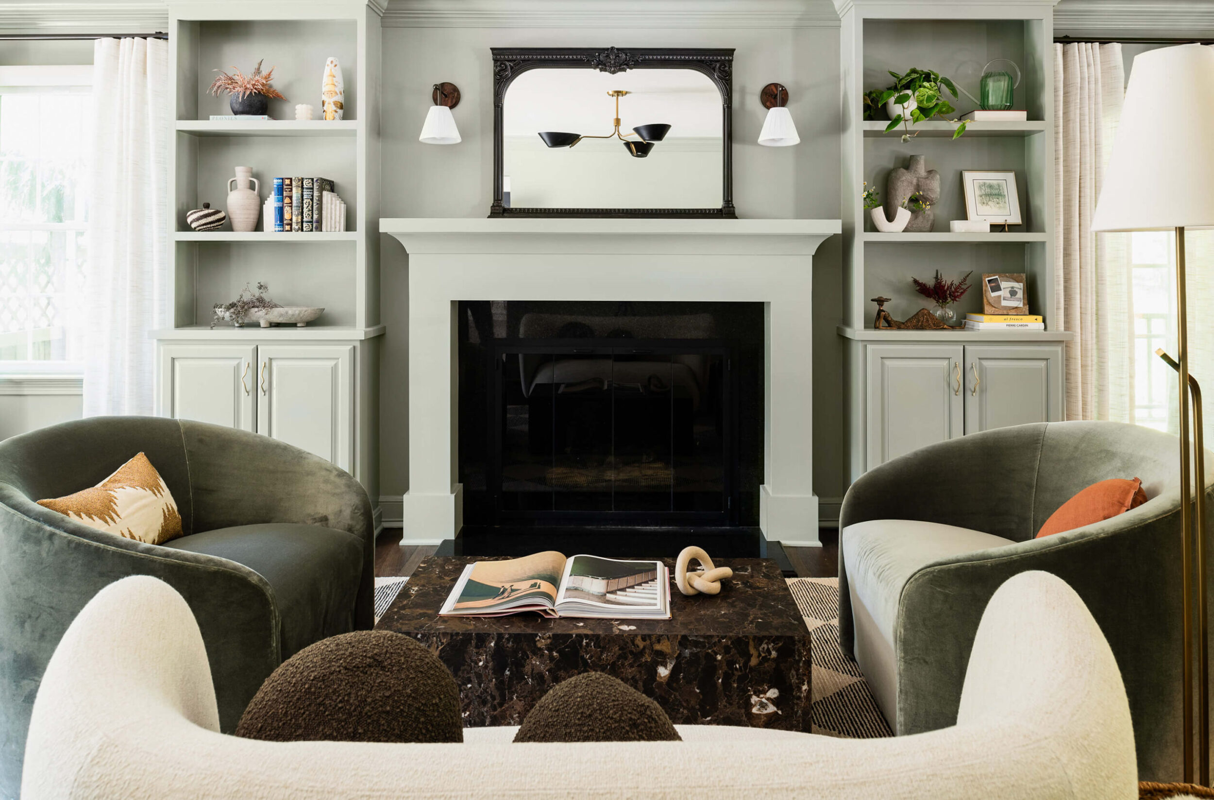 Bright living room with sage built-in shelving, large black fireplace, round velvet chairs, and decorative modern accents.