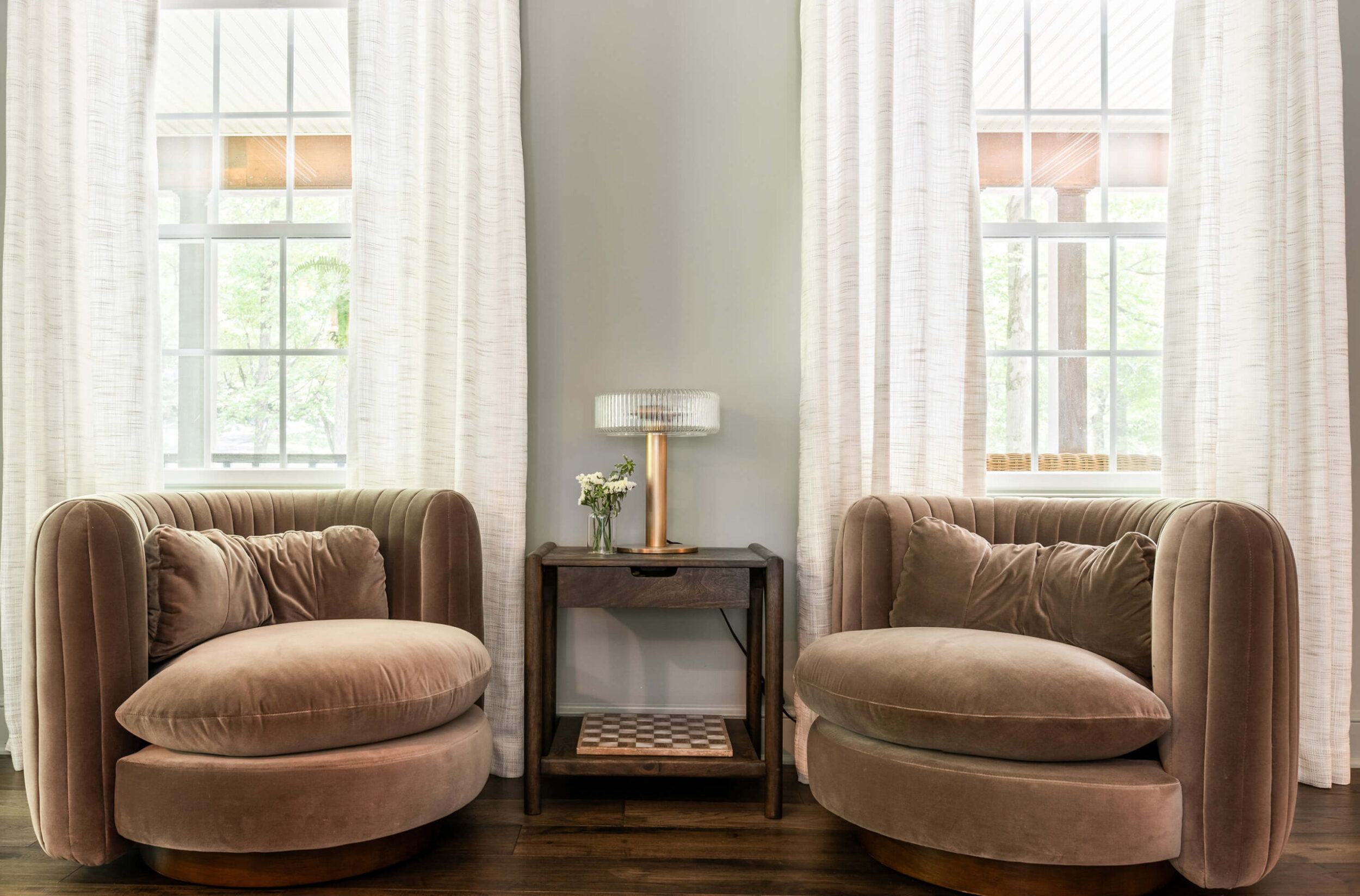 Cozy seating area with plush taupe swivel chairs, side table with lamp and flowers, framed by tall windows with white drapes.