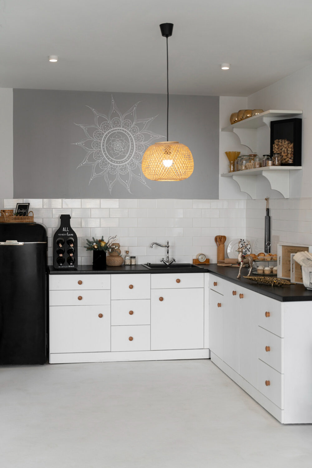 Modern kitchen with white cabinets, black countertop, open shelves, hanging rattan lamp, and decorative wall art.